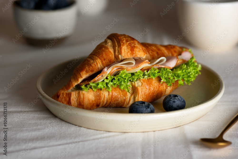 Croissant sandwich with ham, cheese and lettuce, with cup of coffee and blueberries on a table. Croissant sandwich with ham, cheese and lettuce, with cup of coffee and blueberries on a table.