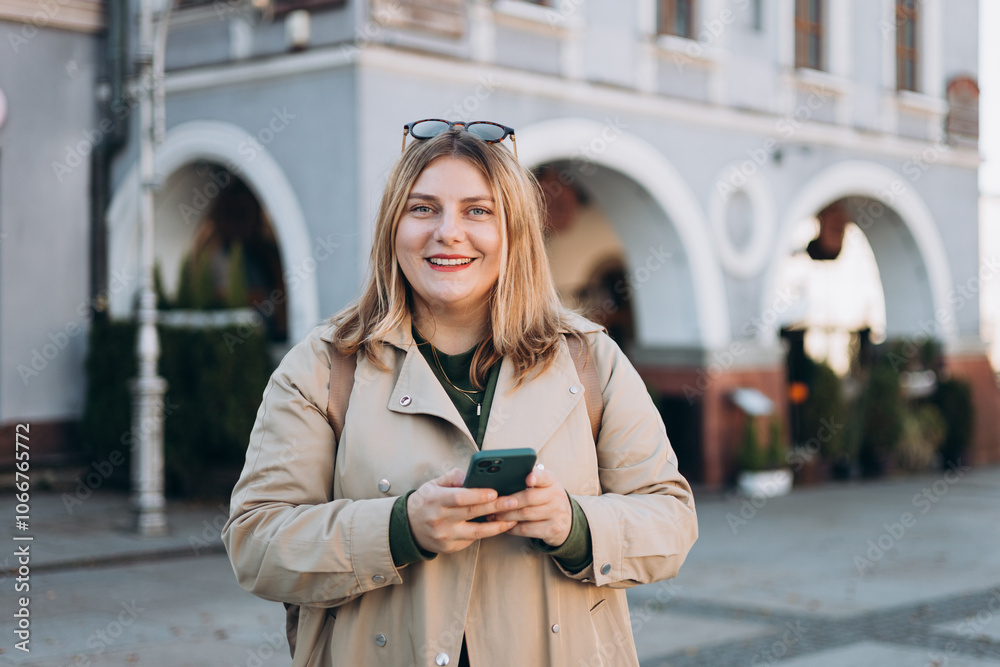 Fototapeta premium Happy cheerful young woman with backpack walking on city street checks her smartphone. Portrait of beautiful 30s girl using smartphone outdoors. Urban lifestyle concept.