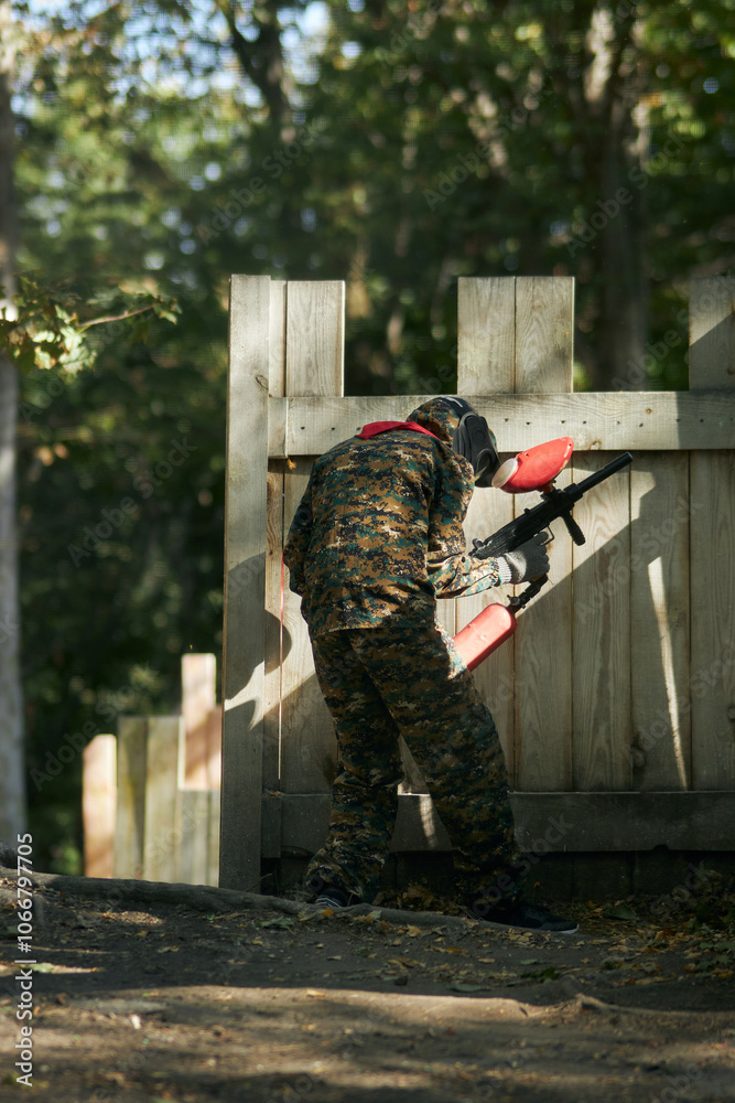 Fototapeta premium Paintball player in protective mask aims gun, takes cover under fire, paint splashes. Paintball player hide behind wooden barricade, shooting paintballs. Paintball players.
