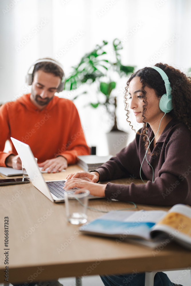 A coworker pair engrossed in their tasks, one taking notes with headphones on while the other types on a laptop, creating an atmosphere of focus and productivity.