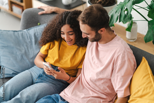 A relaxed couple lying back on a couch, looking at a smartphone, surrounded by plants and bright decor, capturing a serene, cozy moment together in a homey living space.