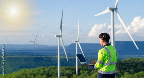 A young engineer is monitoring and inspecting a wind turbine in the field using a digital tablet, checking its performance, with morning sunlight.