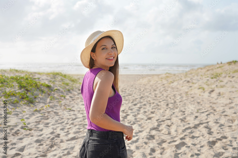 Portrait of young woman with purple tank top and straw hat turns around walking on the sand beach