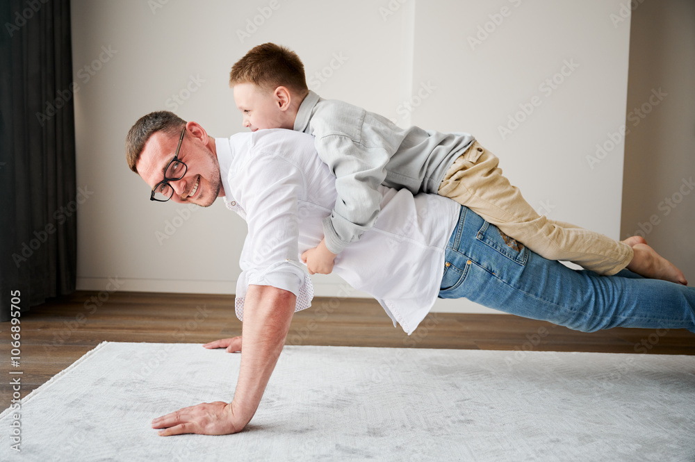 Energetic father and his little son training at home. Kid climbed dad's back while he doing plank. Young family enjoying activity games at home. Energetic father and his little son training at home. Kid climbed dad's back while he doing plank. Young family enjoying activity games at home.