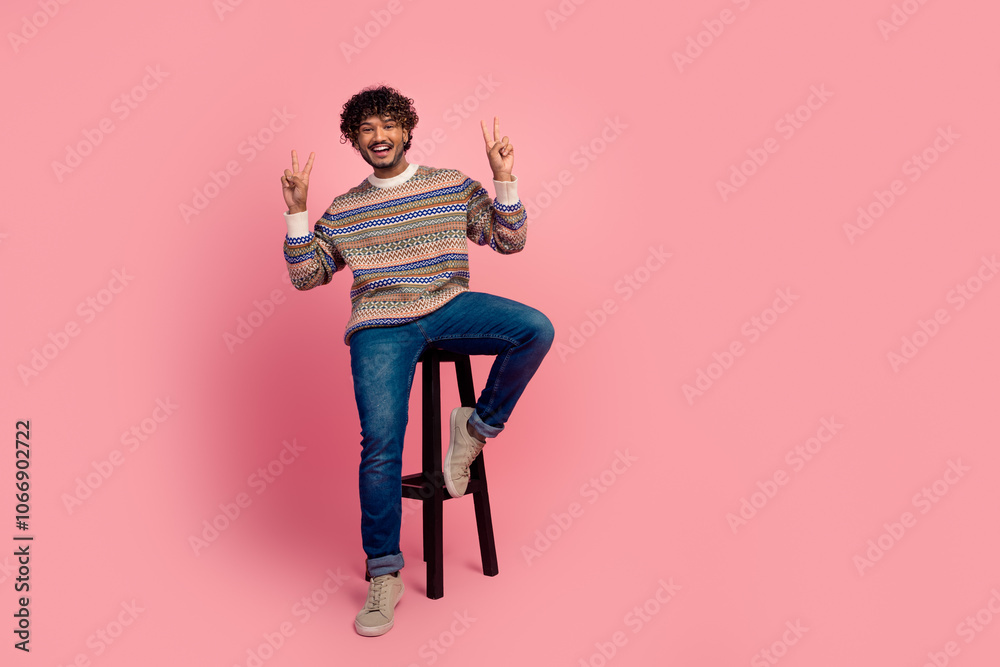 Full size portrait of nice young man sit stool show v-sign isolated on pink color background Full size portrait of nice young man sit stool show v-sign isolated on pink color background
