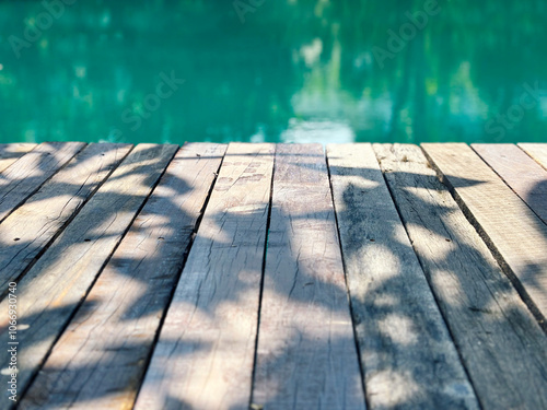 Space on a vintage wooden plank dock or table with the shade of a tree on the floor, the background of the turquoise pond under sunlight in the morning.