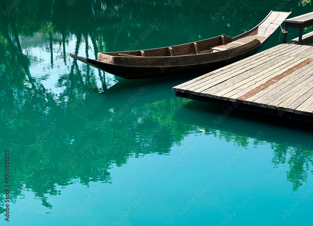 Obraz premium A beautiful Asian-style wooden boat and wooden dock are in the turquoise lake under the morning sunlight. Trees reflect on the water's surface, and there is space for text.