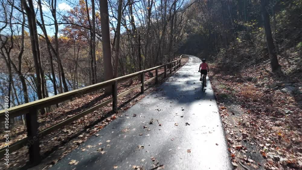 A cyclist rides along a paved trail through a wooded area. The trees are bare, and the ground is covered in fallen leaves. The sun is shining through the branches on C&O Canal. A cyclist rides along a paved trail through a wooded area. The trees are bare, and the ground is covered in fallen leaves. The sun is shining through the branches on C&O Canal.