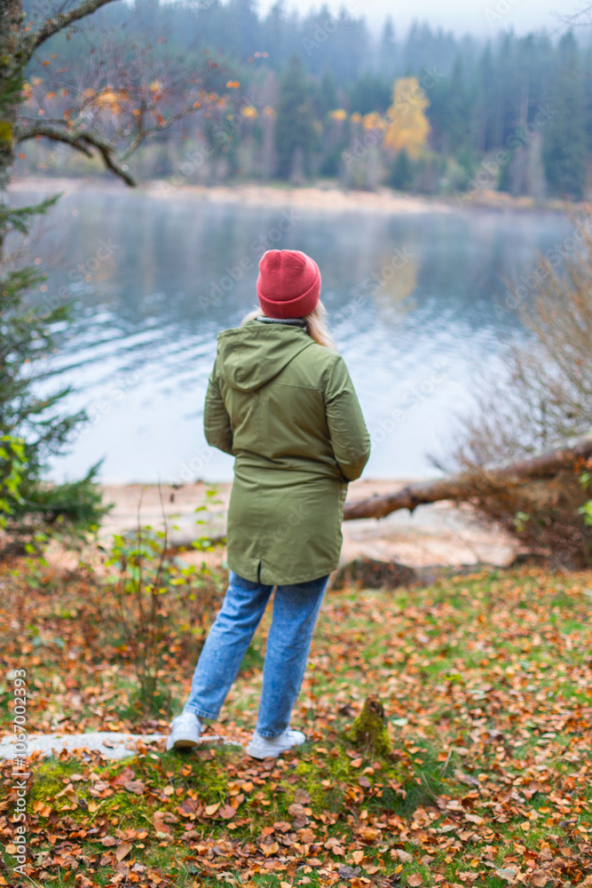 middle-aged caucasian woman in hat and jacket is standing is standing in autumn forest near a lake middle-aged caucasian woman in hat and jacket is standing is standing in autumn forest near a lake