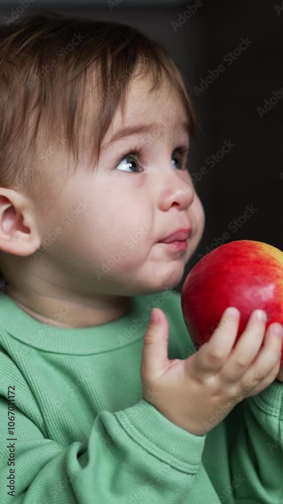 Lovely Caucasian baby boy trying to bite big red apple. Adorable boy in light green shirt looks up turning the fruit in his hands. Close up. Blurred backdrop. Vertical video Lovely Caucasian baby boy trying to bite big red apple. Adorable boy in light green shirt looks up turning the fruit in his hands. Close up. Blurred backdrop. Vertical video