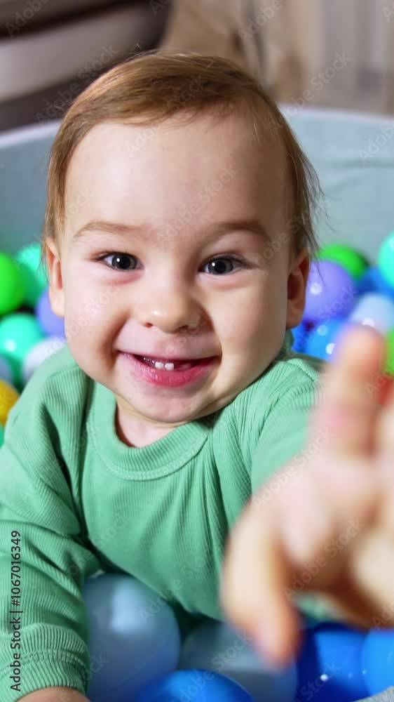 Joyful happy toddler smiling broadly stands in soft basin filled with colorful balls. Cheerful kid trying to reach the camera. Close up. Vertical video