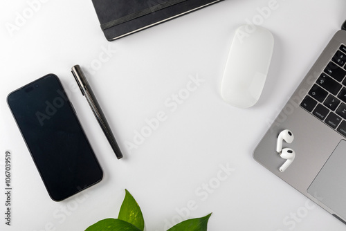 A modern workspace featuring a smartphone, pen, notebook, wireless mouse, laptop, earbuds, and a green plant on a white desk, representing tech-savvy efficiency.
