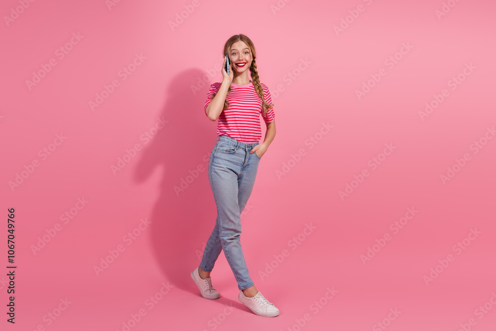 Charming young woman with braids and red lipstick stands against a pink background in casual striped t-shirt, smiling and talking on phone. Charming young woman with braids and red lipstick stands against a pink background in casual striped t-shirt, smiling and talking on phone.