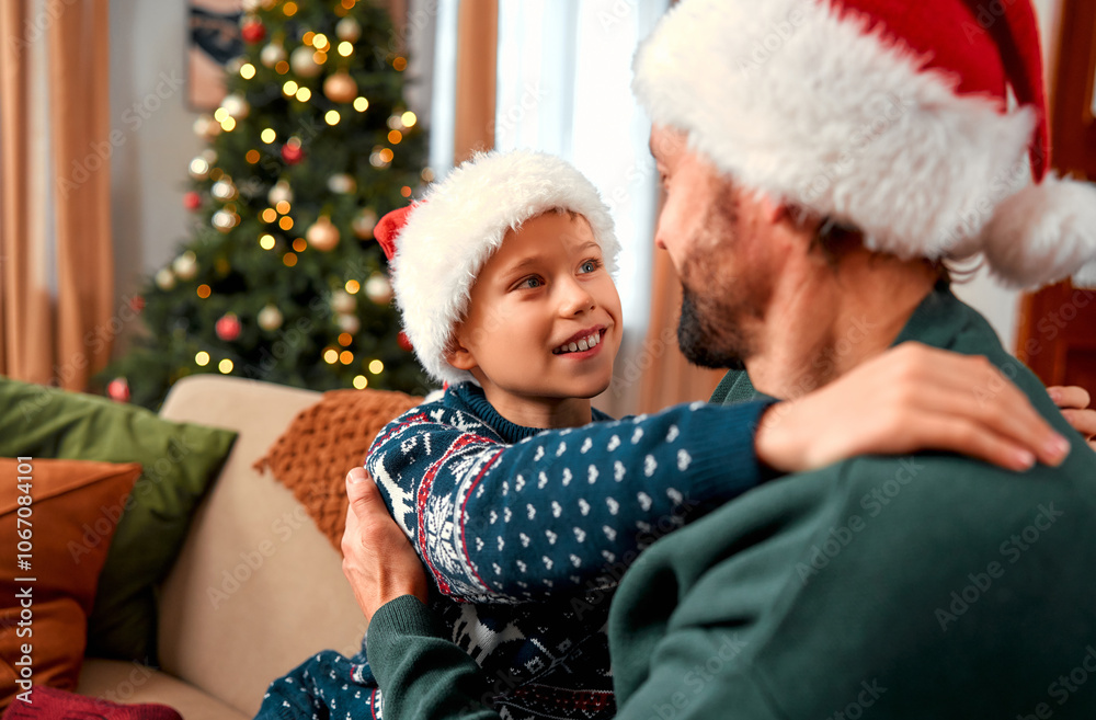Dad and son are sitting together in a cozy, Christmas-decorated living room at home. The boy, wearing a Christmas sweater, hugs his dad. It’s a special time together on Christmas. Dad and son are sitting together in a cozy, Christmas-decorated living room at home. The boy, wearing a Christmas sweater, hugs his dad. It’s a special time together on Christmas.