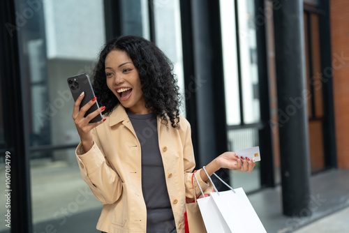 Happy young woman holding a smartphone and credit card, shopping online while carrying shopping bags in an urban environment.