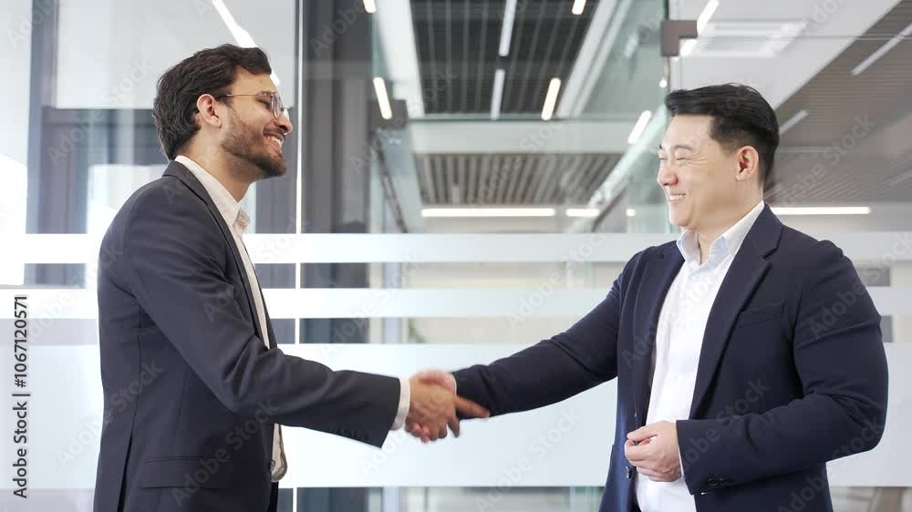 Two businessmen in formal suits shaking hands after deal while standing in a modern business office. Male coworkers smile, suggesting successful collaboration, professional relationship and teamwork Two businessmen in formal suits shaking hands after deal while standing in a modern business office. Male coworkers smile, suggesting successful collaboration, professional relationship and teamwork