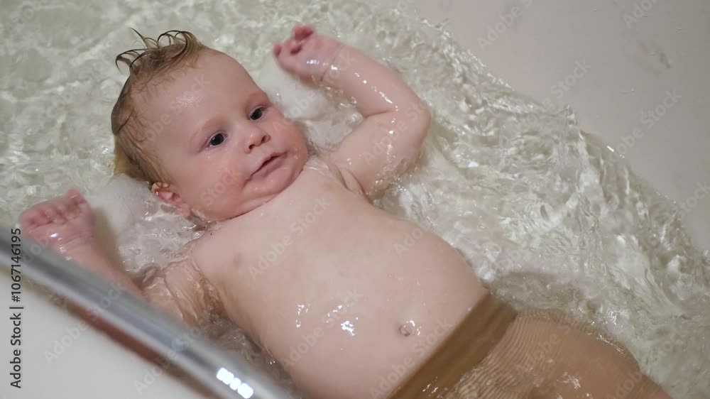Little kid is waving his arms and splashing, happily relaxing on soft towel. Cheerful infant baby is happily bathing in bathtub filled with water, enjoying delightful bath