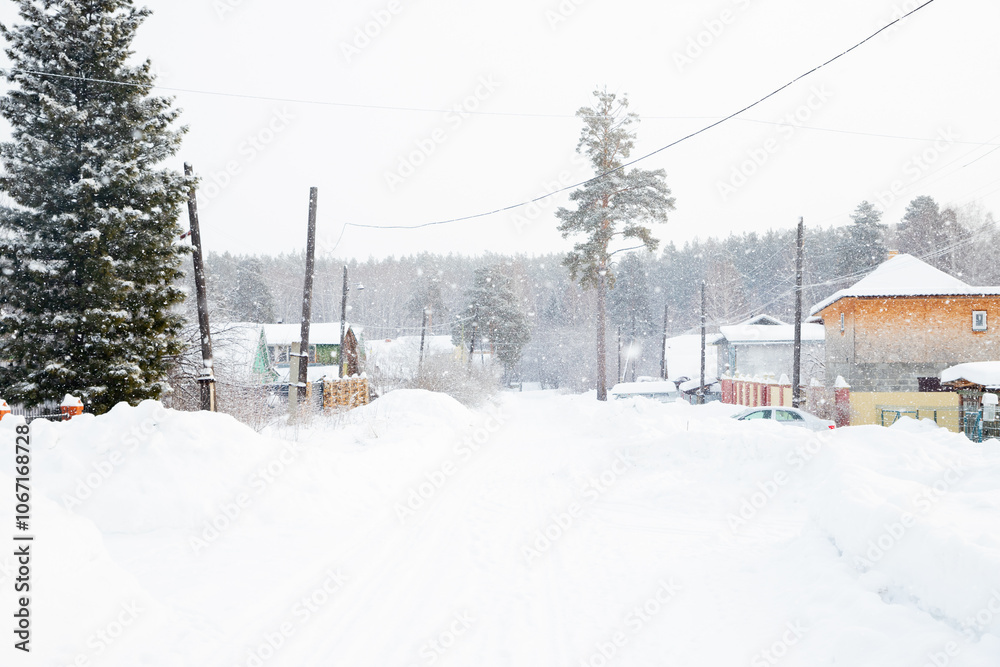Snow blankets the village street in a quiet rural areas, with homes and tall trees peeking through the falling snowflakes on a winter day.
