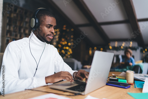 African American man working remotely on laptop, wearing headphones and focused. Cozy workspace with warm lighting, festive decorations. Background shows relaxed atmosphere and blurred holiday lights photo