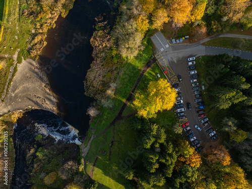 Top-down aerial photo of the Jagala River and Jägala Waterfall in Estonia. On the left the river flows with dark, almost black water speckled with white foam. On the right parking area with many cars