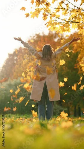 Joyful woman resting in park in autumn day, throwing yellow leaves in air, rear view, vertical video. Happiness and joy in fall season, sunny weather and good mood, full-length with female person