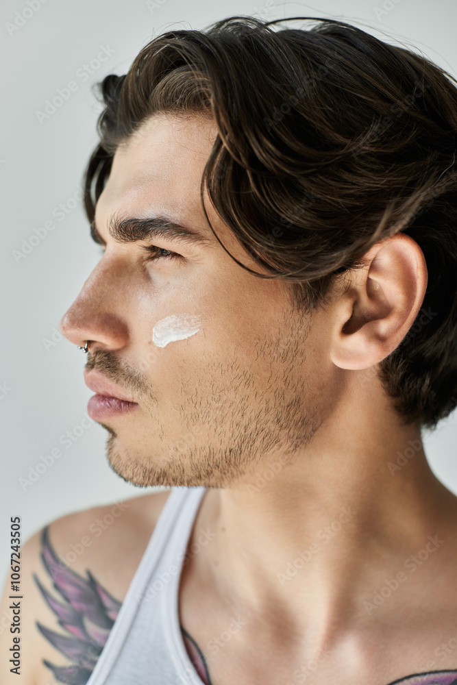 Fototapeta premium A stylish man in a tank top carefully applies cream to his face, revealing his unique tattoos and focused expression.