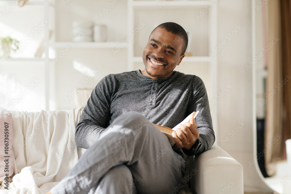 Smiling smart african american guy reading book, sit on chair, enjoy comfort and free time in white living room interior. Relax, hobby and literatura at home. High quality photo Smiling smart african american guy reading book, sit on chair, enjoy comfort and free time in white living room interior. Relax, hobby and literatura at home. High quality photo