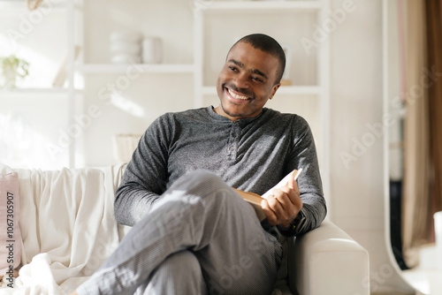 Smiling smart african american guy reading book, sit on chair, enjoy comfort and free time in white living room interior. Relax, hobby and literatura at home. High quality photo