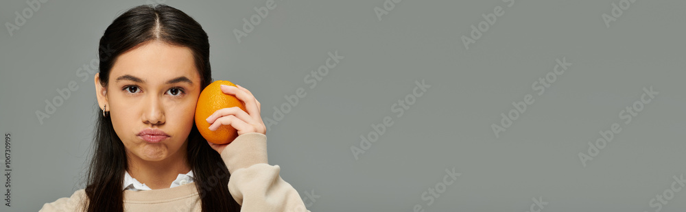 A young woman in fashionable attire holds an orange to her cheek, lost in thought and emotion. A young woman in fashionable attire holds an orange to her cheek, lost in thought and emotion.