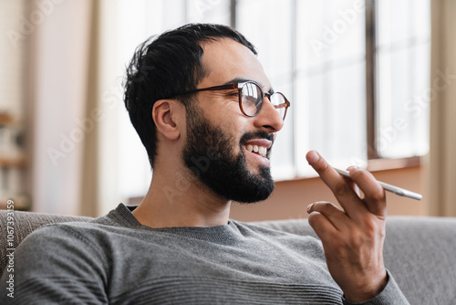 Positive handsome young Arabian business man talking on speaker on cellphone, recording voice message, looking away, thinking, activating modern smart home application photo