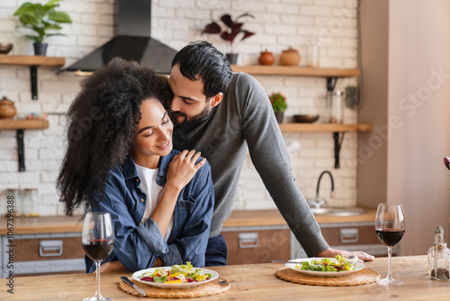 I love you. Cheerful young arabian man husband is hugging his african wife woman with fondness while standing behind her. Lady is sitting at table in kitchen and smiling. Love relationship family photo