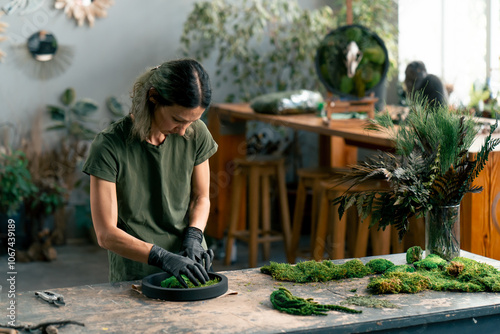 in large green workshop a thin woman with green hair in black gloves at a large table collects a composition from stabilized moss photo