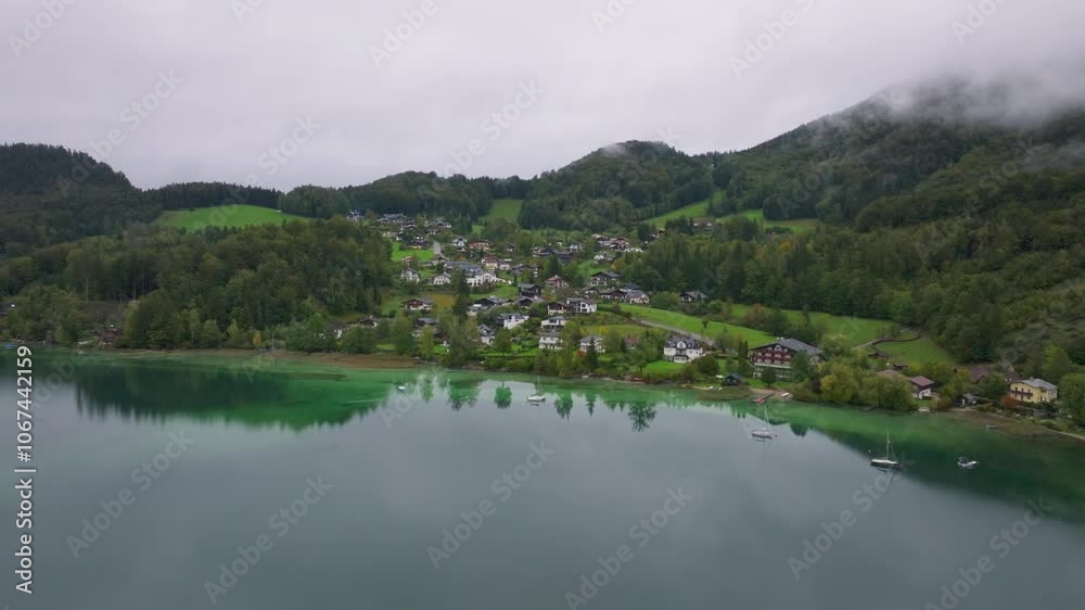 Camera is panning over a turquoise lake in the Alps with a small village on its shore. Green Alps mountains and low clouds are in the background Camera is panning over a turquoise lake in the Alps with a small village on its shore. Green Alps mountains and low clouds are in the background
