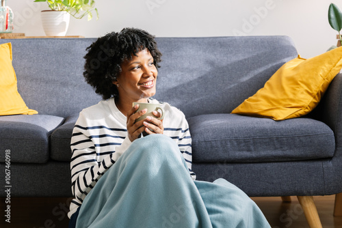 girl sitting in living room with cup of hot coffee enjoying under blanket. Joyful black girl sitting on floor by the sofa wrapped in blanket in living room with cup of hot coffee photo