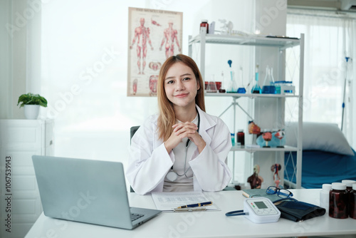 A woman in a white lab coat sits at a desk with a laptop open in front of her