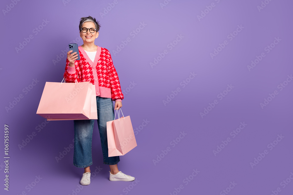 Full size photo of pretty retired female hold gadget shopping bags wear trendy red outfit isolated on purple color background Full size photo of pretty retired female hold gadget shopping bags wear trendy red outfit isolated on purple color background