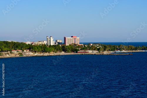 View of Antalya's coastline with modern buildings and lush greenery along the Mediterranean Sea, under a clear blue sky. 