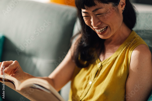 Foto Relaxed asian woman reading book on couch in bright living room.