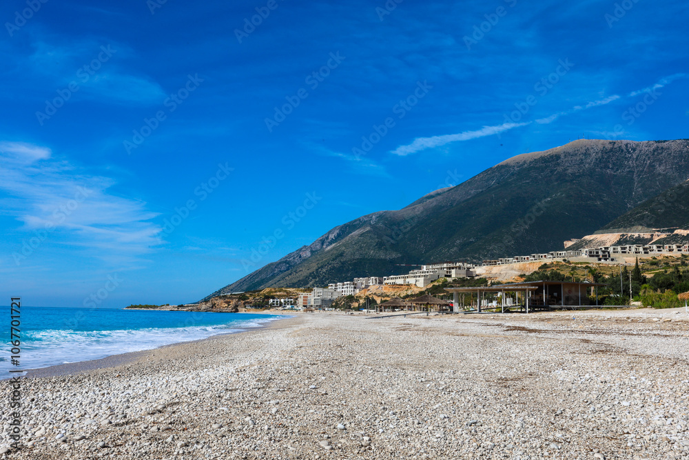 Fototapeta premium wide pebble beach of Palasës on the Albanian Riviera, deserted outside the tourist season, in the background an apartment complex being built, the Logar Pass and the hilltops of the Logar National Par