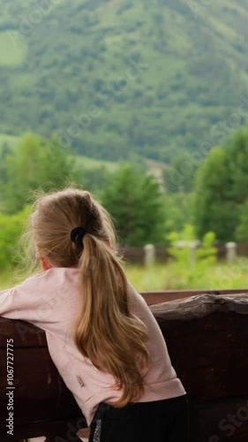 Curious little girl looks at flying up helicopter leaning on large wooden log railing on terrace of rural cafe on cloudy summer day slow motion photo