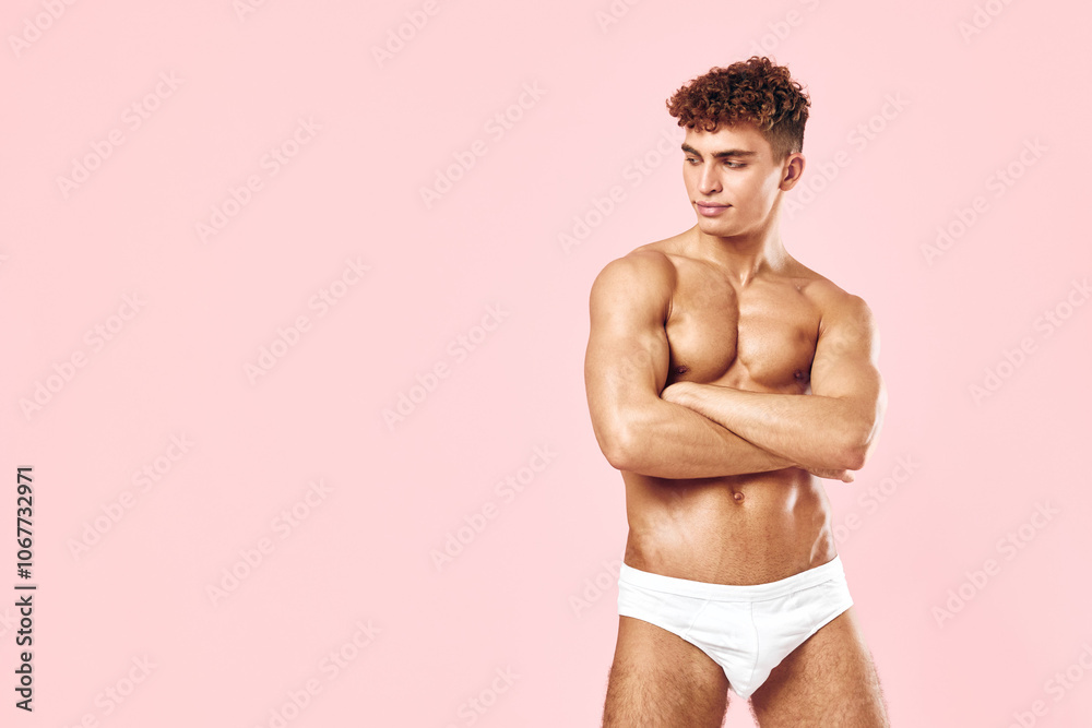 Fototapeta premium Confident young man posing with crossed arms, showcasing a toned physique against a soft pink background. Body positivity and fitness concept.