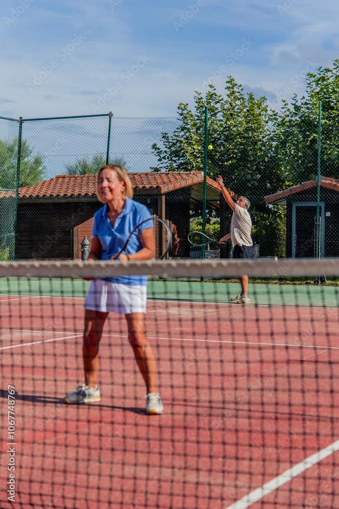 vertical Senior tennis couple playing match at club with woman in foreground as husband serves vertical Senior tennis couple playing match at club with woman in foreground as husband serves
