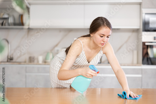 Young woman in dress cleaning kitchen. Positive housewife with rag and detergent spray polish cleans dining table from grease and dirt