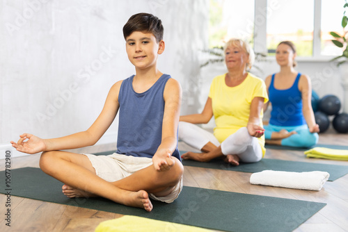 Mother, grandmother and adolescence son do exercise half lotus position of Ardha Padmasana Sidhasana with hands clasped in mudra knowledge pose. Concept of communication of generations, joint leisure