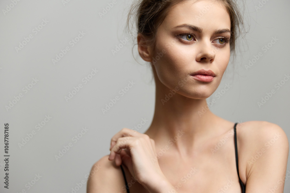 Naklejka premium Elegant young woman with natural makeup, gazing thoughtfully, dressed in a simple black top. Soft gray background emphasizes her serene expression and beauty.