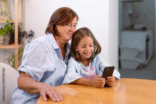 Smiling girl uses smartphone with her grandmother at table, sharing joyful and engaging moment of technology and bonding in warmth of their home 