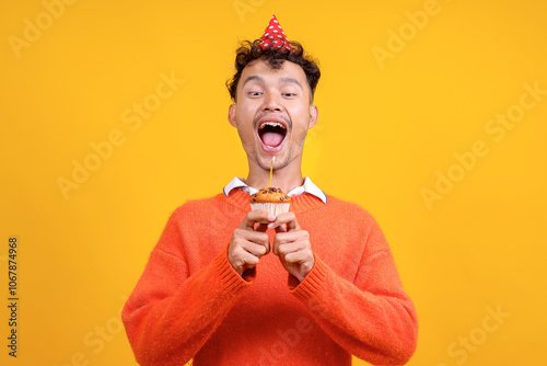 Portrait of Excited Young Asian Man Open Mouth Ready to Blowing Candle on Cupcake Isolated on Yellow Background