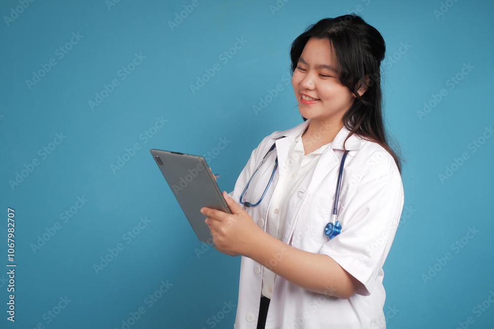 Asian young female doctor in white lab coat working on digital tablet and smiling while standing isolated on blue background Asian young female doctor in white lab coat working on digital tablet and smiling while standing isolated on blue background