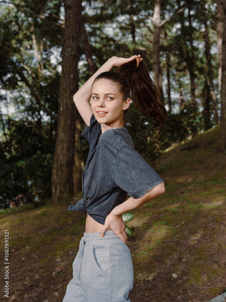 Young woman with long hair casually tying it back while standing in a forest, wearing a relaxed outfit and smiling Outdoor lifestyle and nature friendly vibe Young woman with long hair casually tying it back while standing in a forest, wearing a relaxed outfit and smiling Outdoor lifestyle and nature friendly vibe