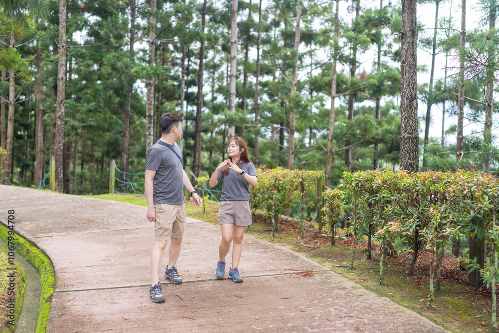 Obraz premium A Chinese-Malaysian couple in their 30s walk together in a green park with abundant water in Kuala Lumpur, Malaysia, on a sunny day in June 2024.